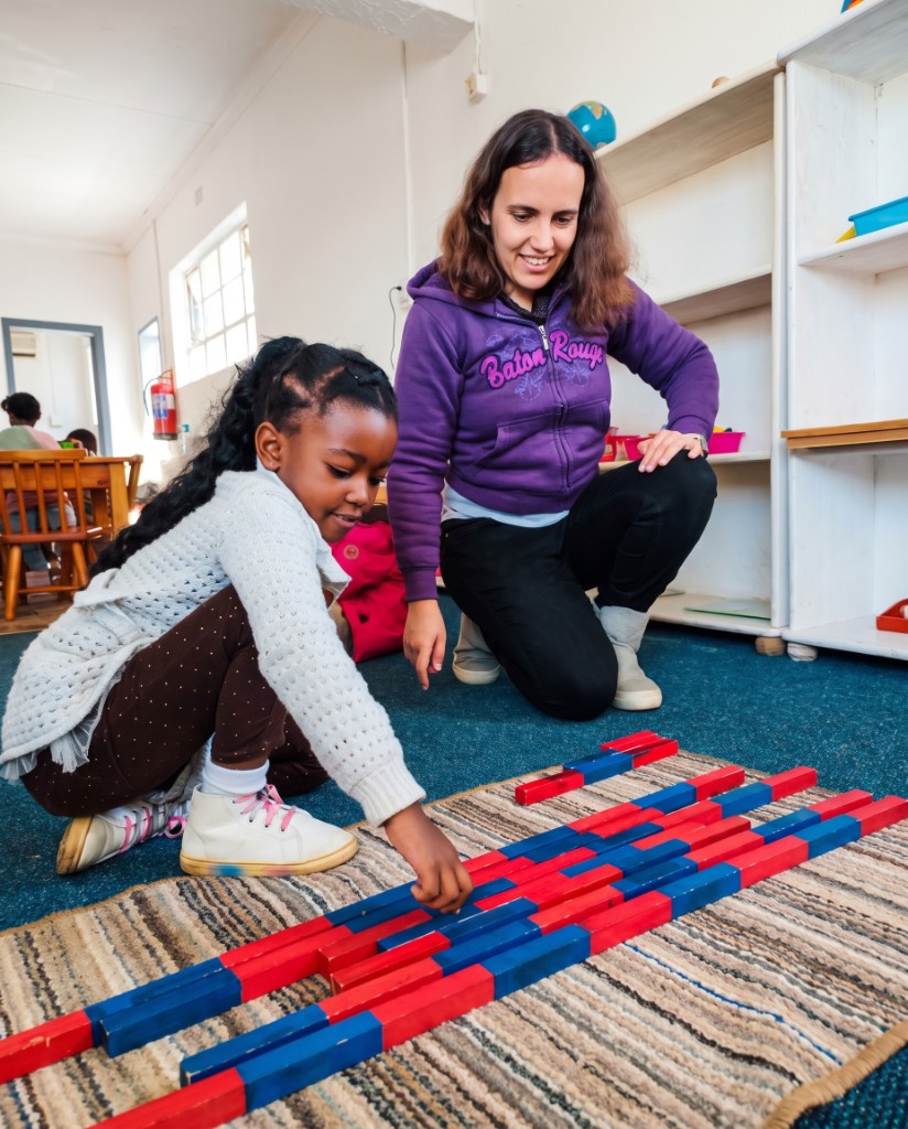 An Addnum ECD educator kneeling beside a young learner arranging Montessori number rods on a classroom rug