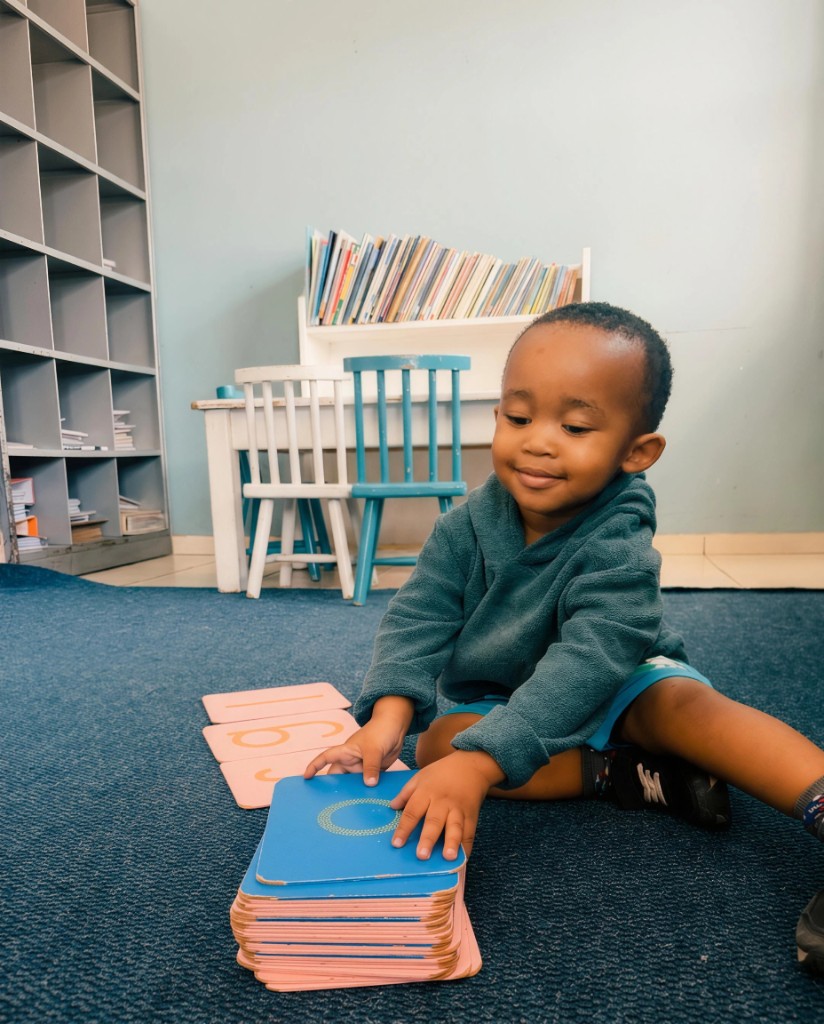 A young toddler sitting on a blue classroom carpet, smiling while arranging large educational cards