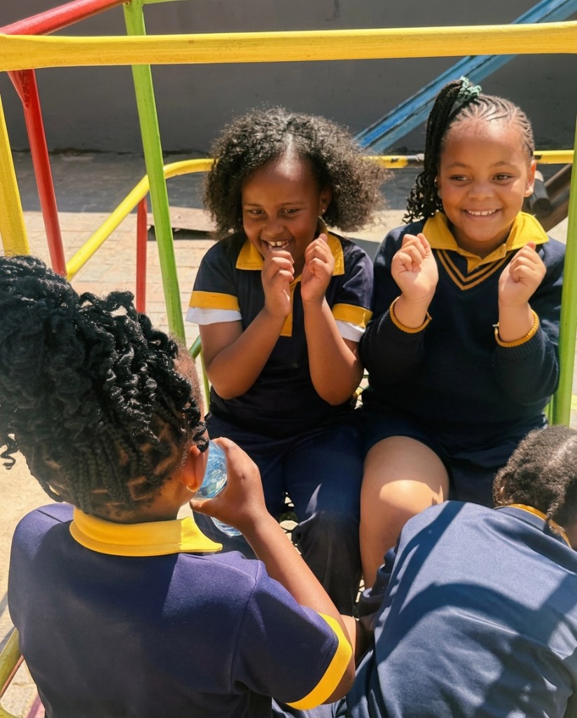 Young Addnum ECD learners in navy and yellow uniforms laughing together on playground equipment