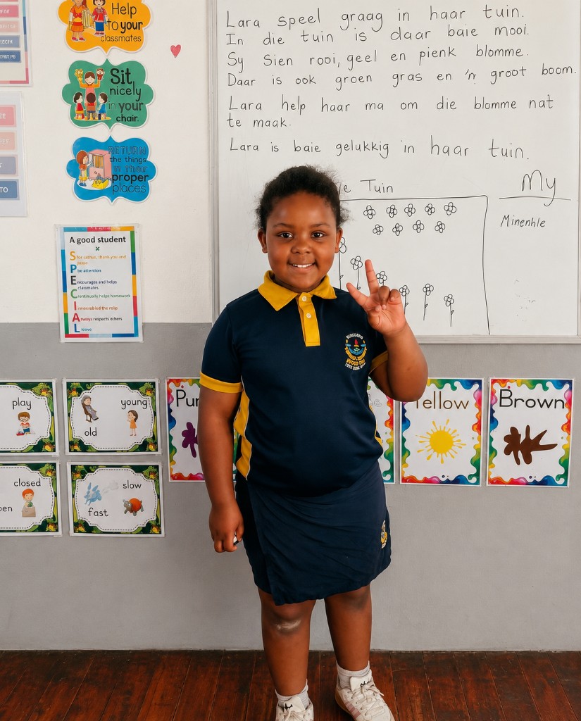 A happy young Addnum ECD learner in navy and yellow uniform smiling in a classroom with a whiteboard behind her